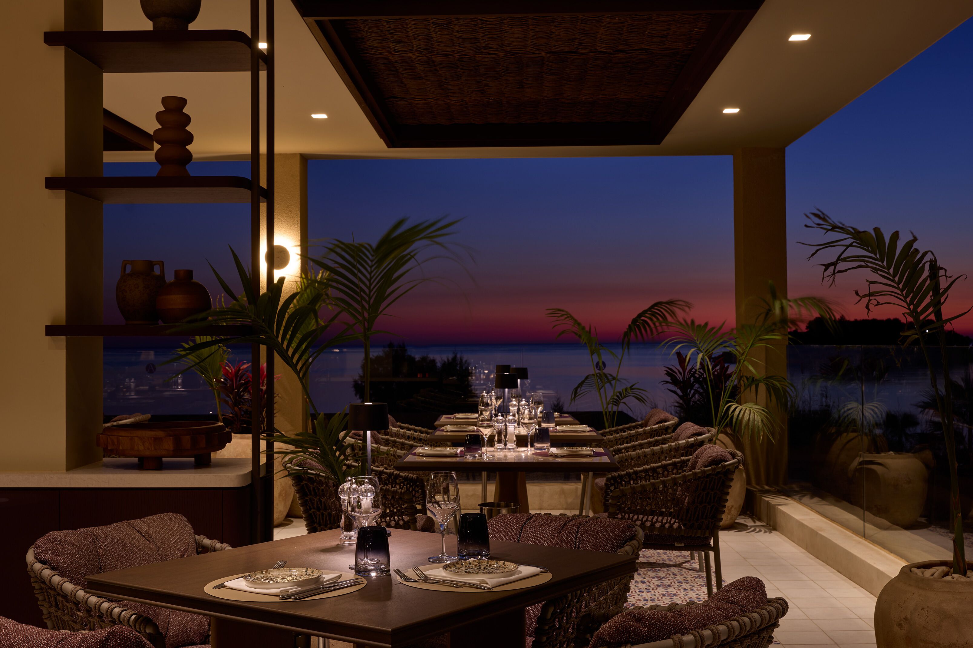 A dining area with a view of the sunset over the ocean, furnished with wicker chairs and a table set for two.
