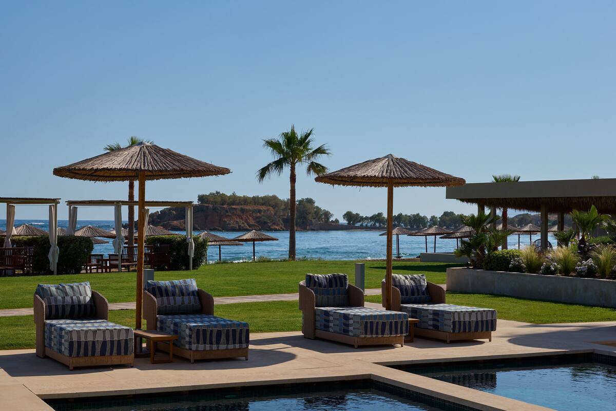 Zona junto a la piscina con sillones de descanso y sombrillas, con vistas a un cuerpo de agua y una costa rocosa.