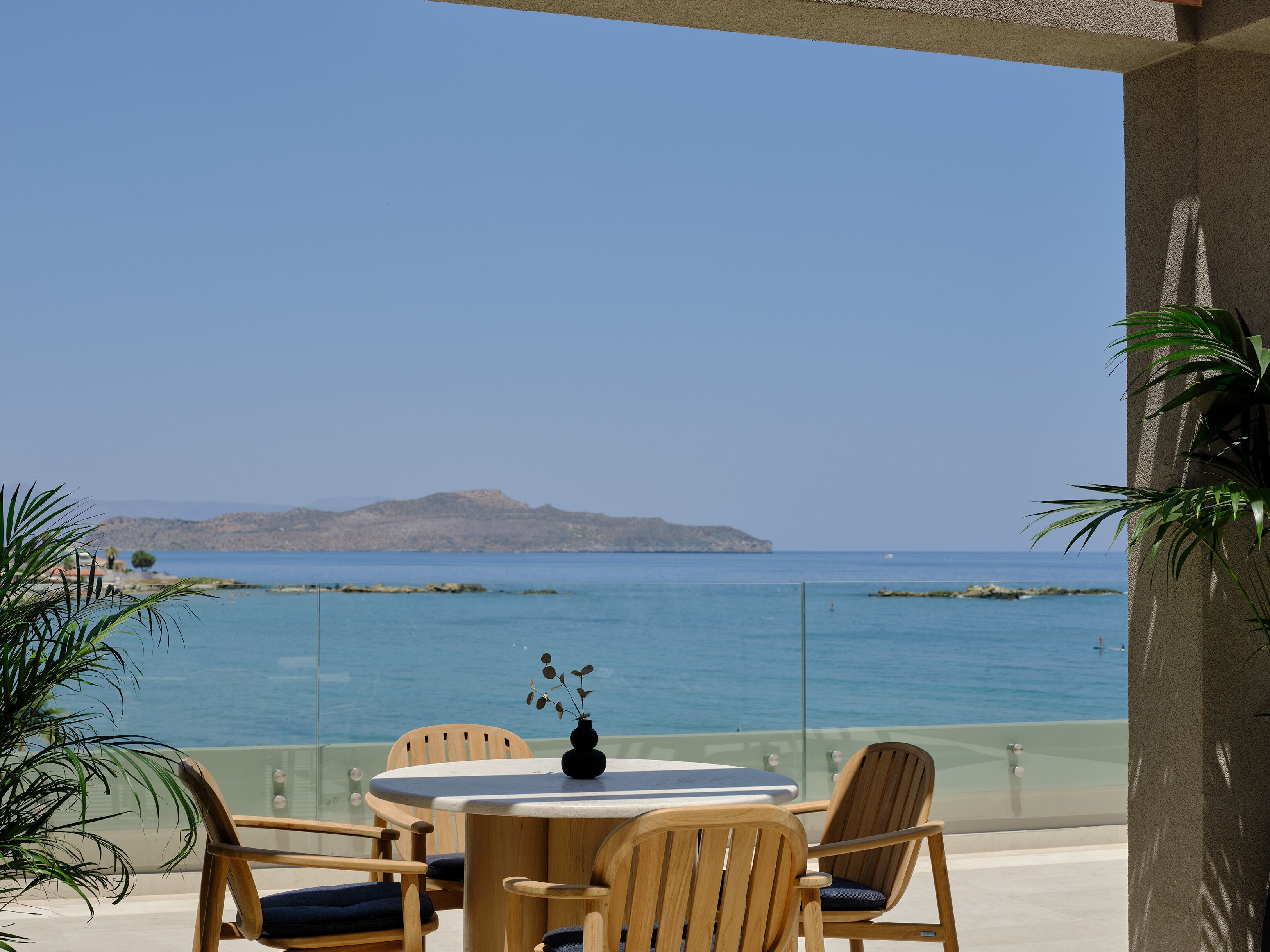 A glass-enclosed outdoor seating area with a table and chairs, overlooking a calm sea and distant hills.