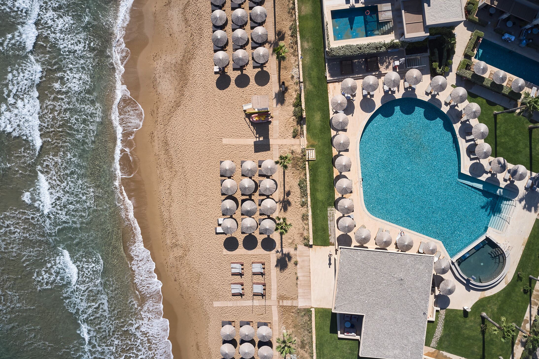 An aerial view of a resort with a large swimming pool, beach area with umbrellas, and lounge chairs.