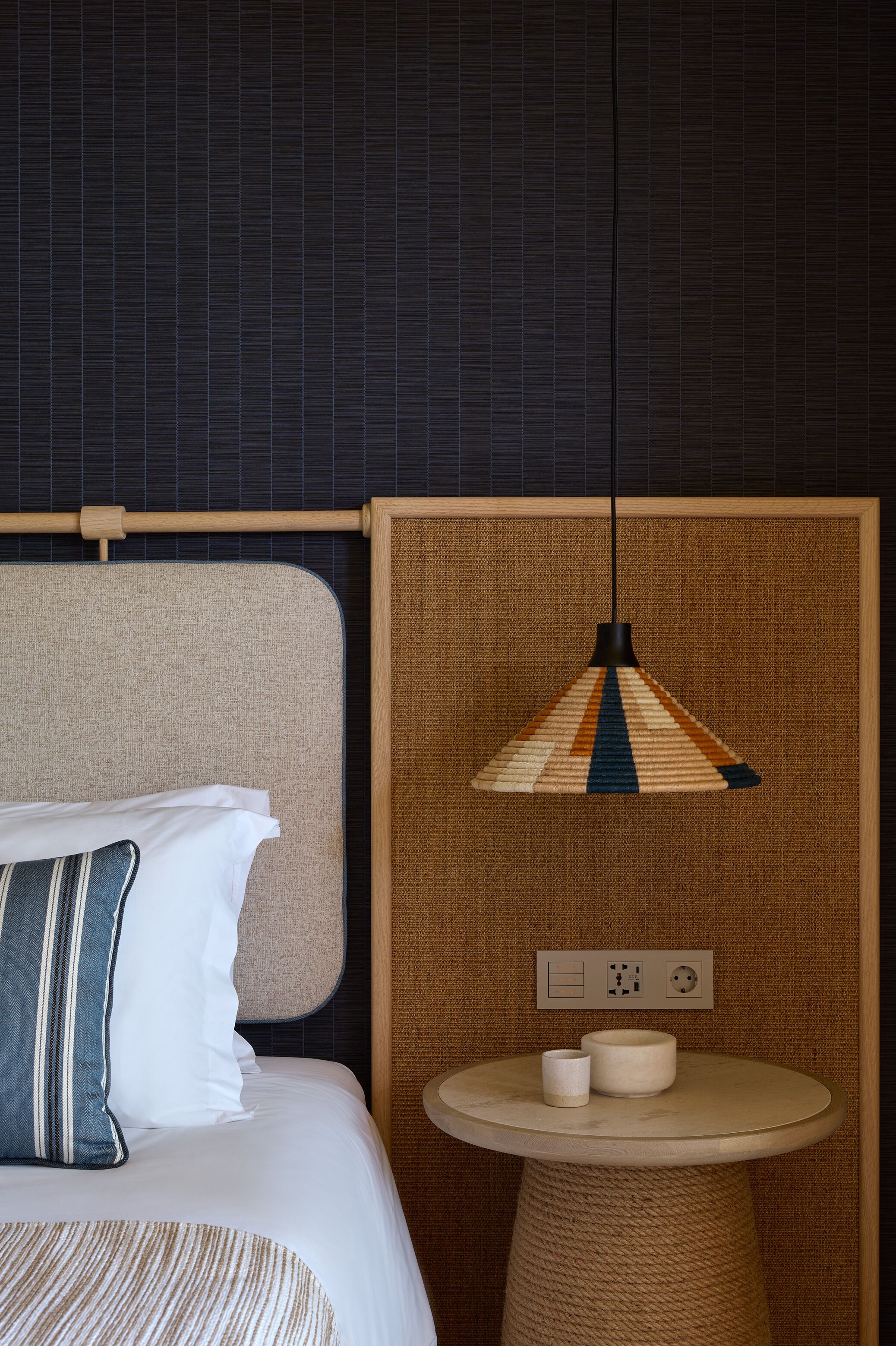 A neatly made bed with a striped pillow, a round table with two cups, and a hanging lamp with a striped shade.