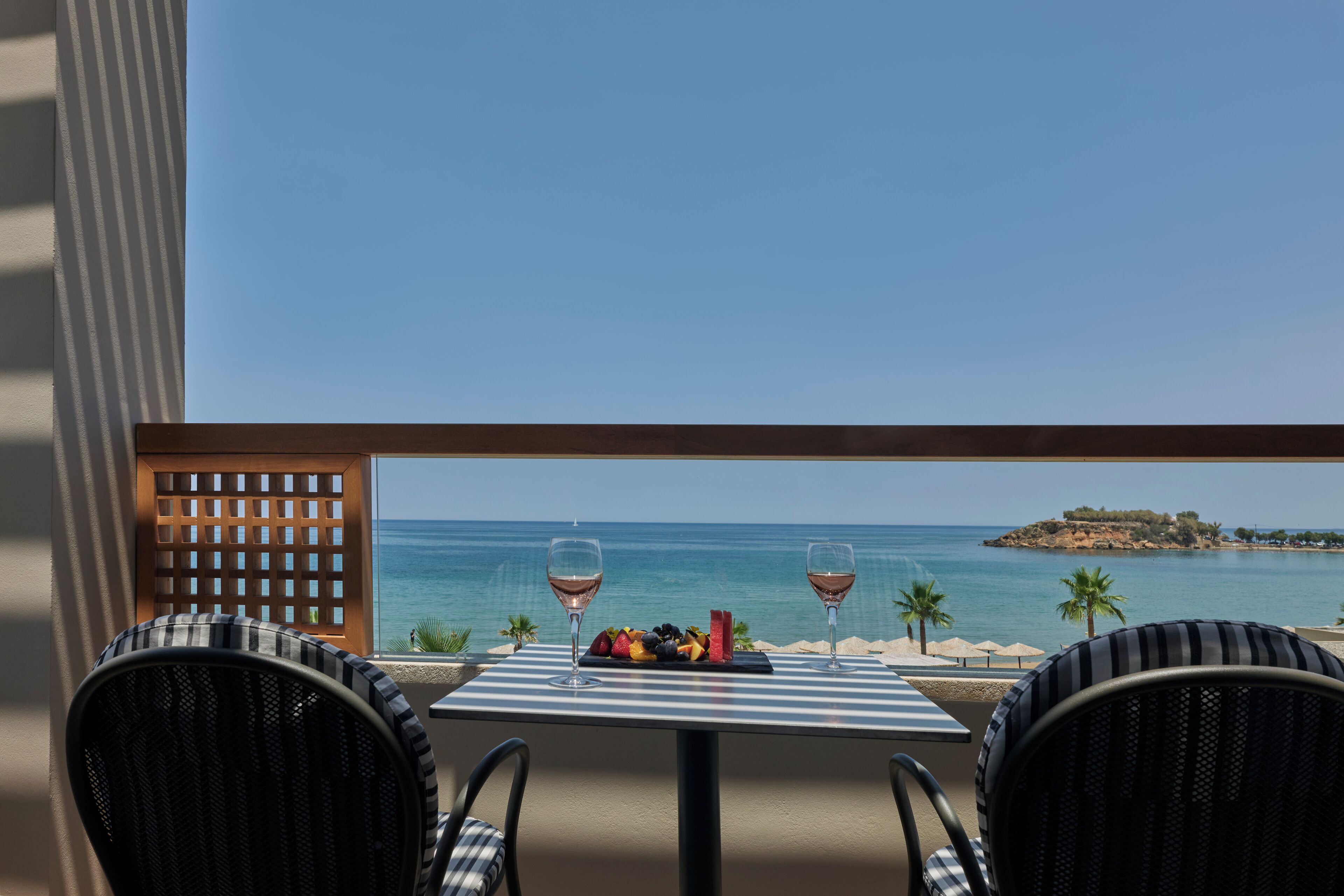 A table with two chairs and a fruit platter, two wine glasses, and a view of the sea and a small island.