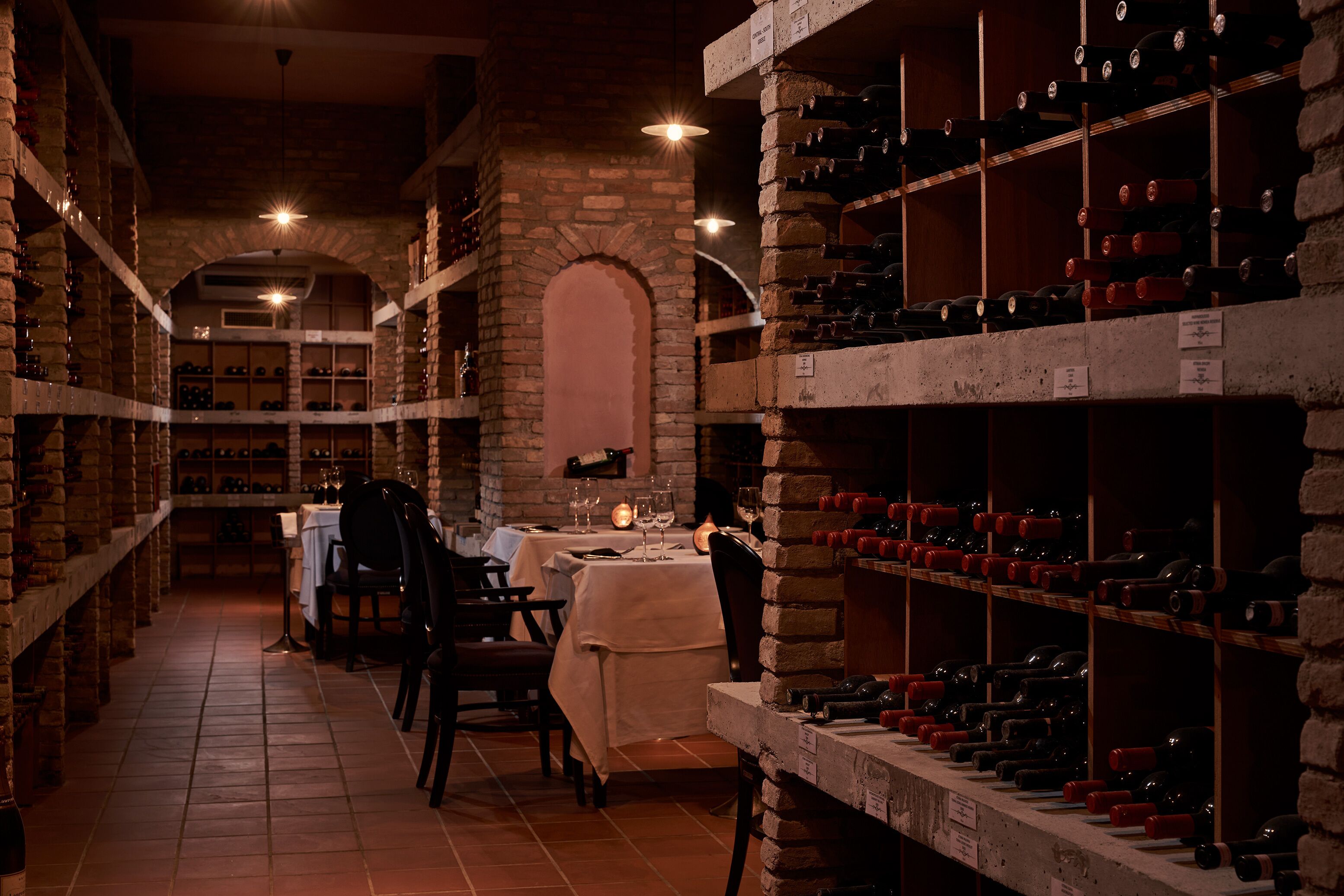 A wine cellar with shelves of wine bottles, a dining area with a table and chairs, and a brick wall with an arched window.