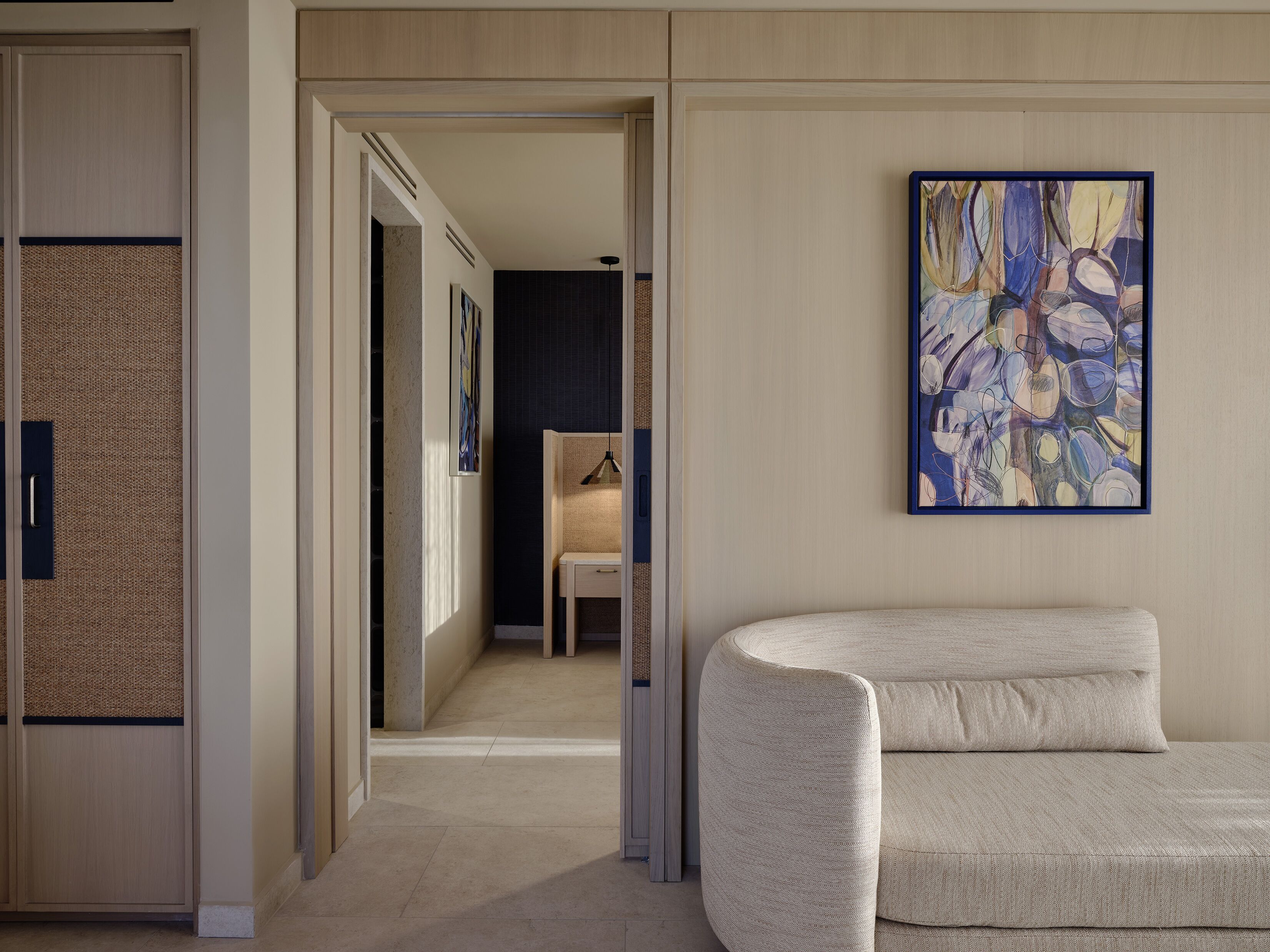 A hotel room with a sofa, a painting, and a view into the hallway.