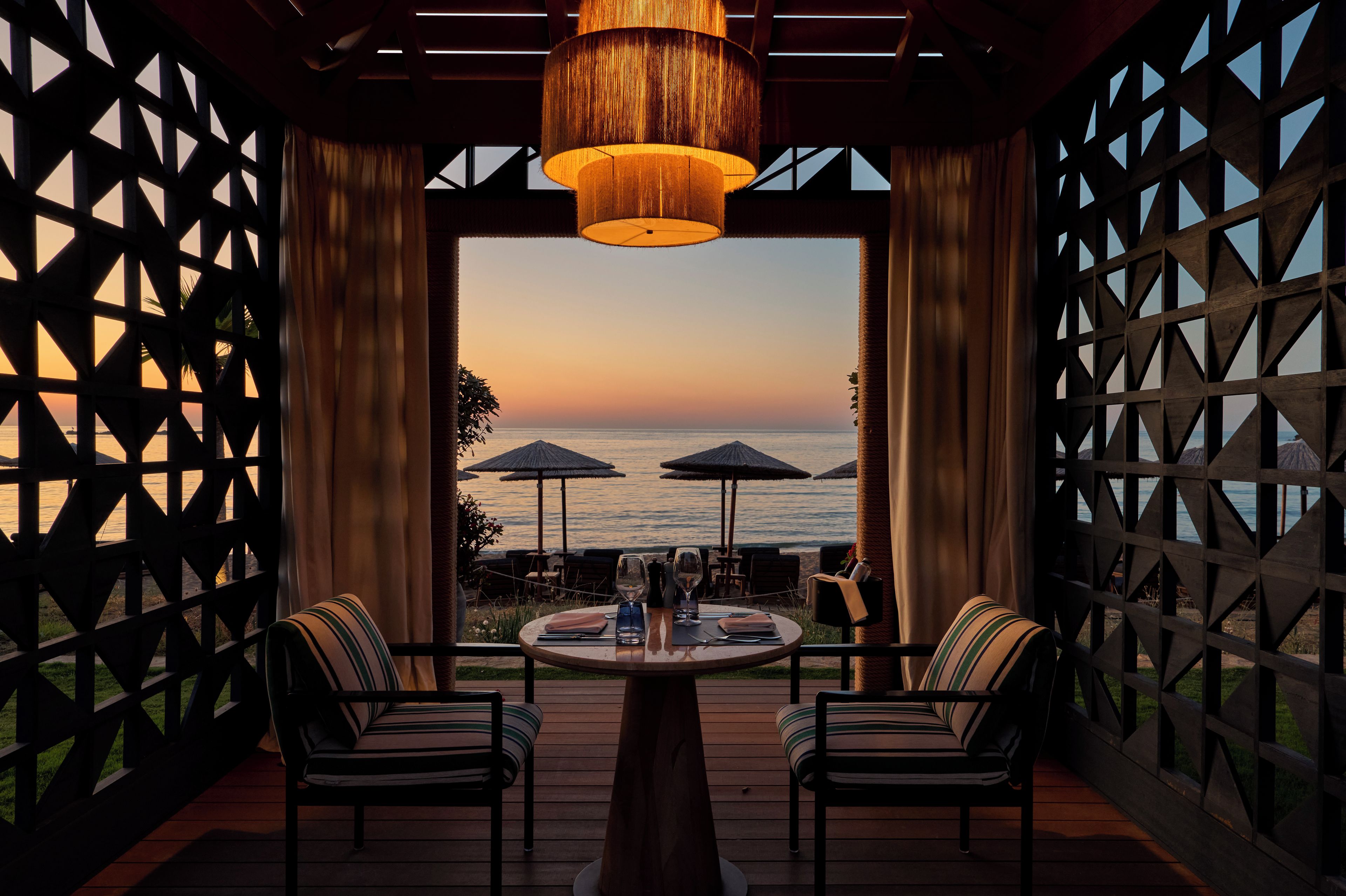 An outdoor dining area with a view of the sea, featuring a table with chairs, a hanging light fixture, and a wooden lattice wall.