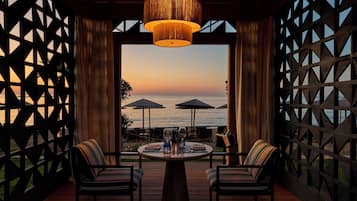 An outdoor dining area with a view of the sea, featuring a table with chairs, a hanging light fixture, and a wooden lattice wall.
