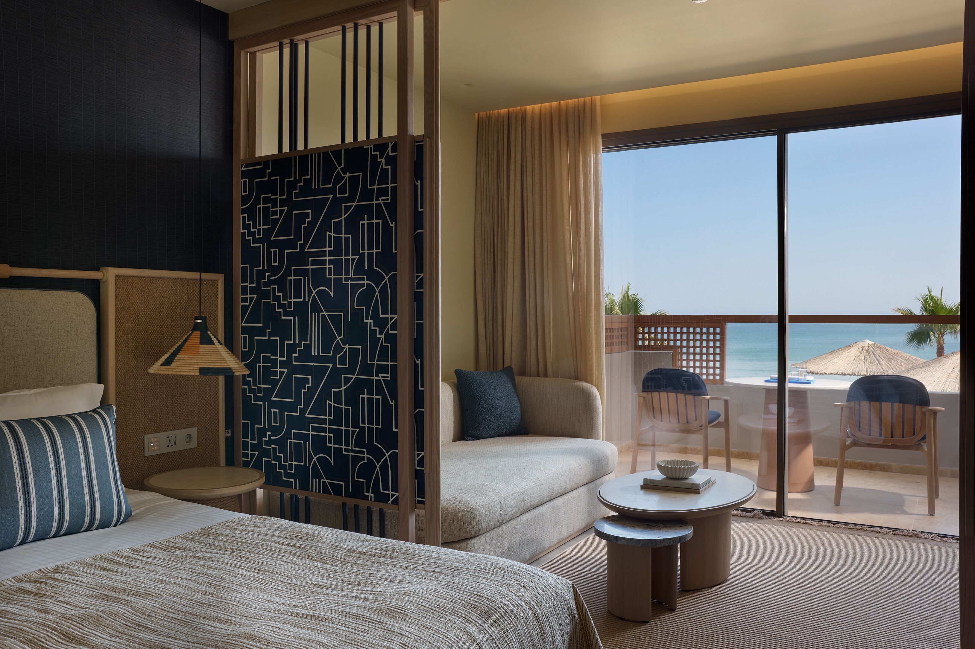A hotel room with a bed, a sofa, a small table, and a balcony with a view of the beach.