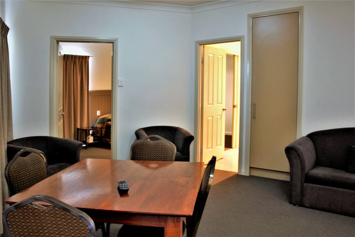 A hotel room with a wooden dining table, four chairs, two sofas, and two doors leading to other rooms.
