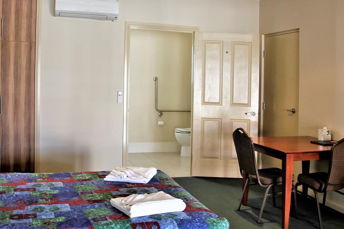 A hotel room with a bed, a table, and a chair. There is a bathroom visible through an open door.