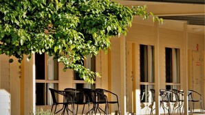 A building with a covered outdoor seating area, multiple windows, and a tree providing shade.