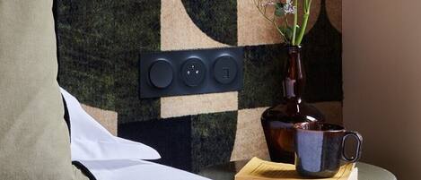 Una habitación con pared de cuadros, un panel eléctrico negro, un florero con flores y una taza sobre una mesita.