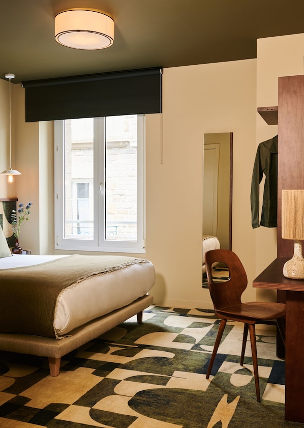 Un dormitorio con cama, sillón, escritorio, lámpara y ventana con vistas al exterior.