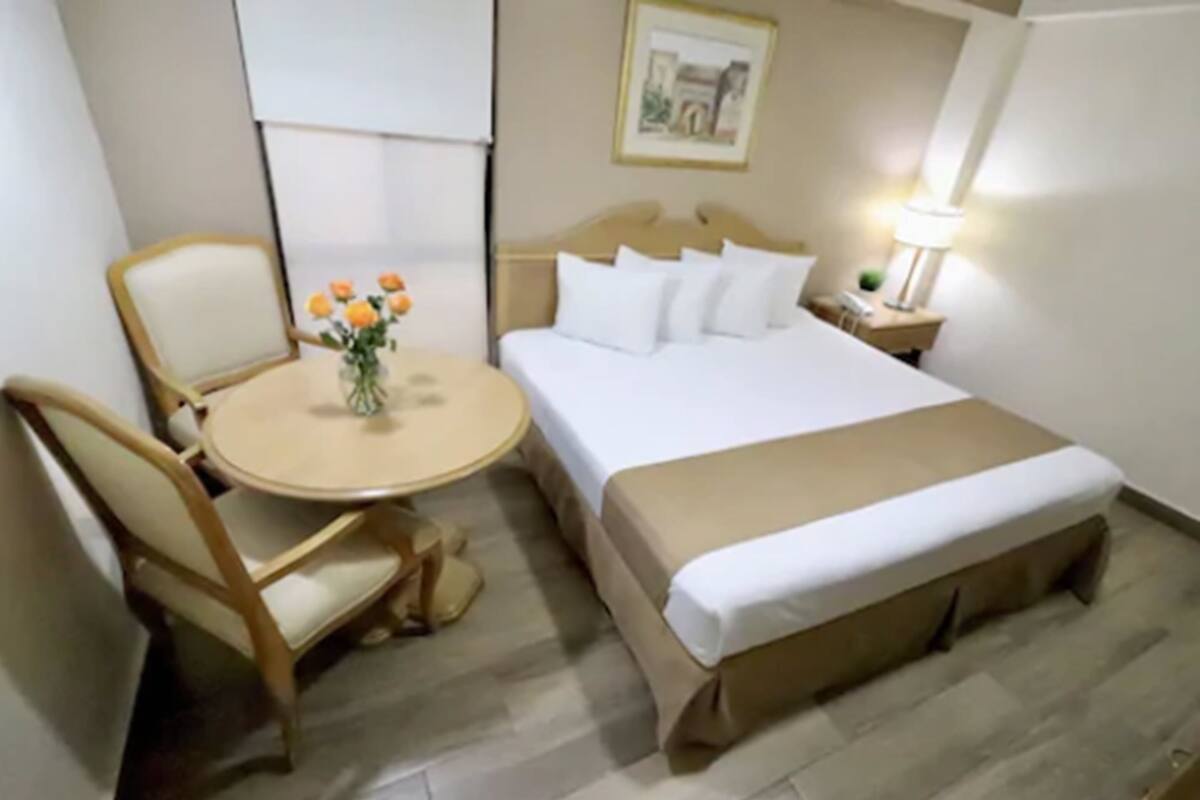 A hotel room with a bed, a bedside table with a lamp, a chair, and a small round table with a vase of flowers.
