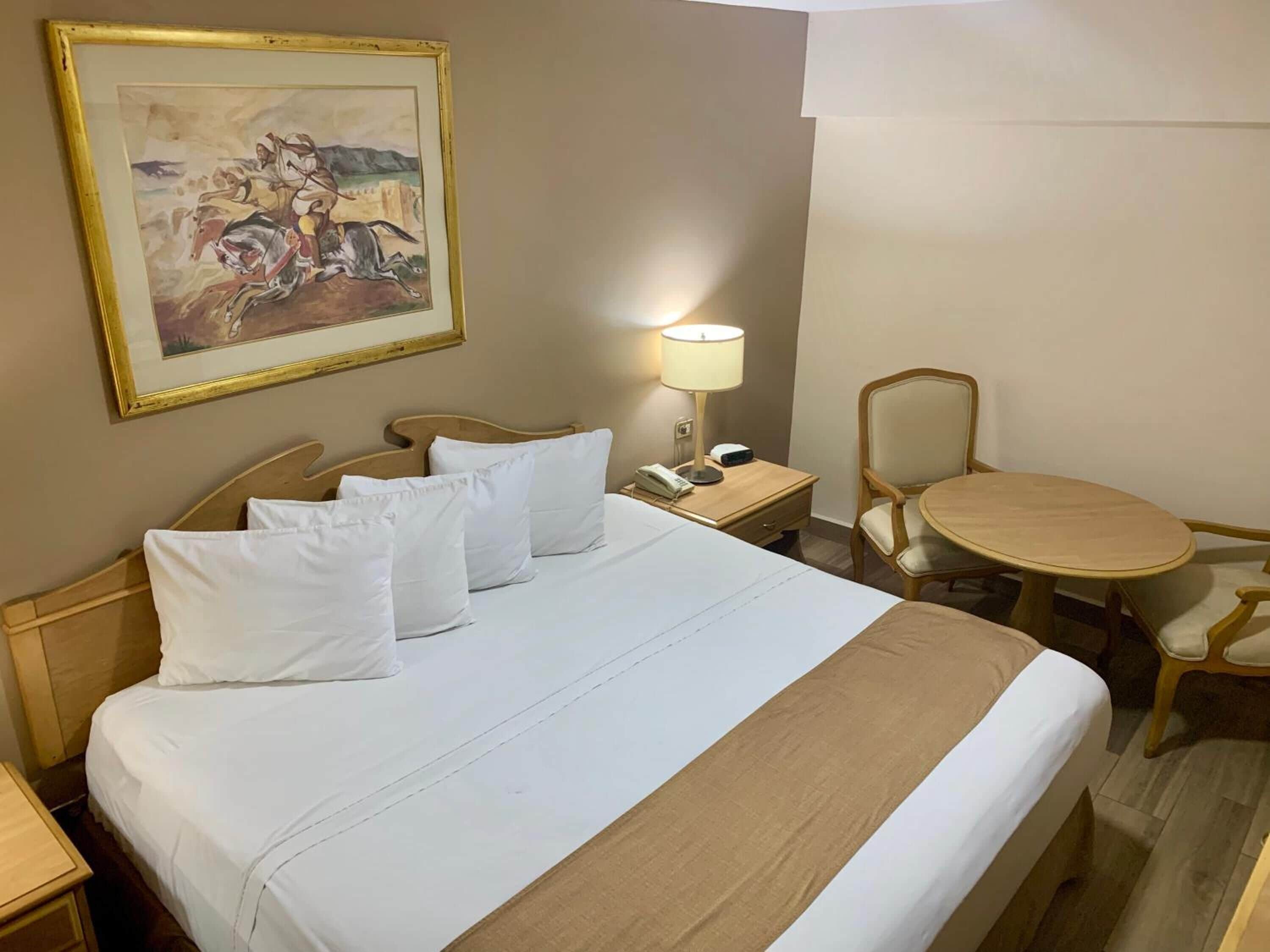 Una habitación de hotel con cama, mesita de noche con lámpara, silla y una mesa de comedor.