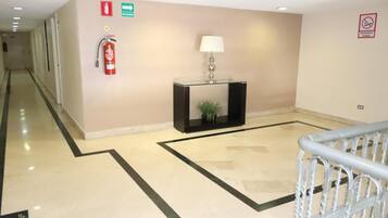 Un vestíbulo de hotel con un extintor, un letrero de salida de emergencia verde, una consola negra con una lámpara y una baranda de metal.