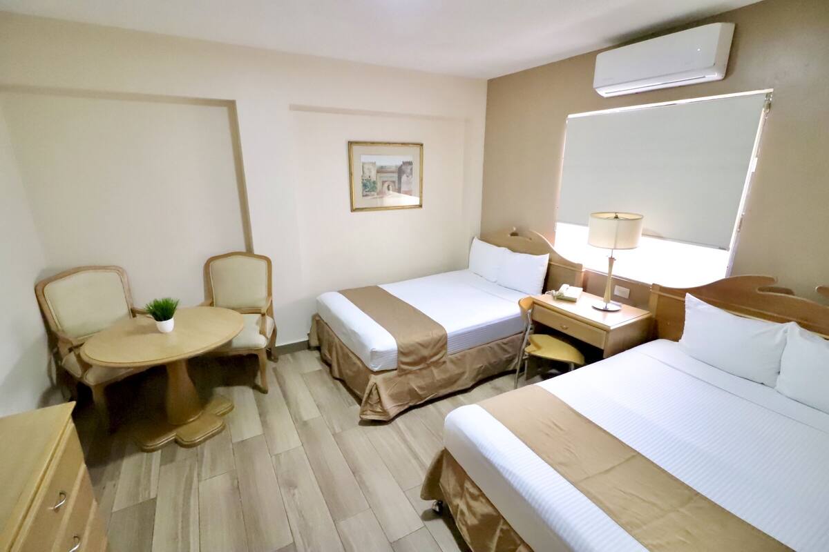 A hotel room with two beds, a desk, a chair, a small table with a plant, and a picture on the wall.