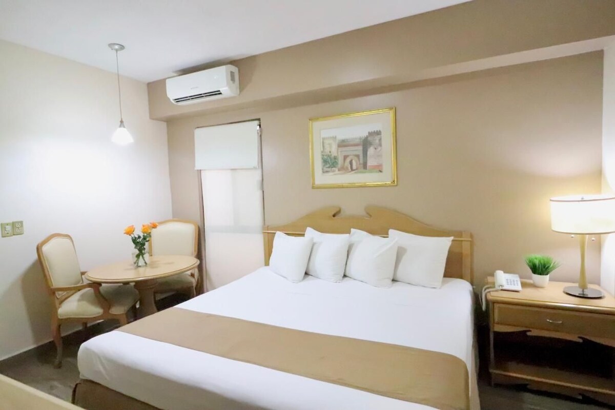 A hotel room with a bed, a bedside table with a lamp, a chair, a small table with flowers, and an air conditioning unit.