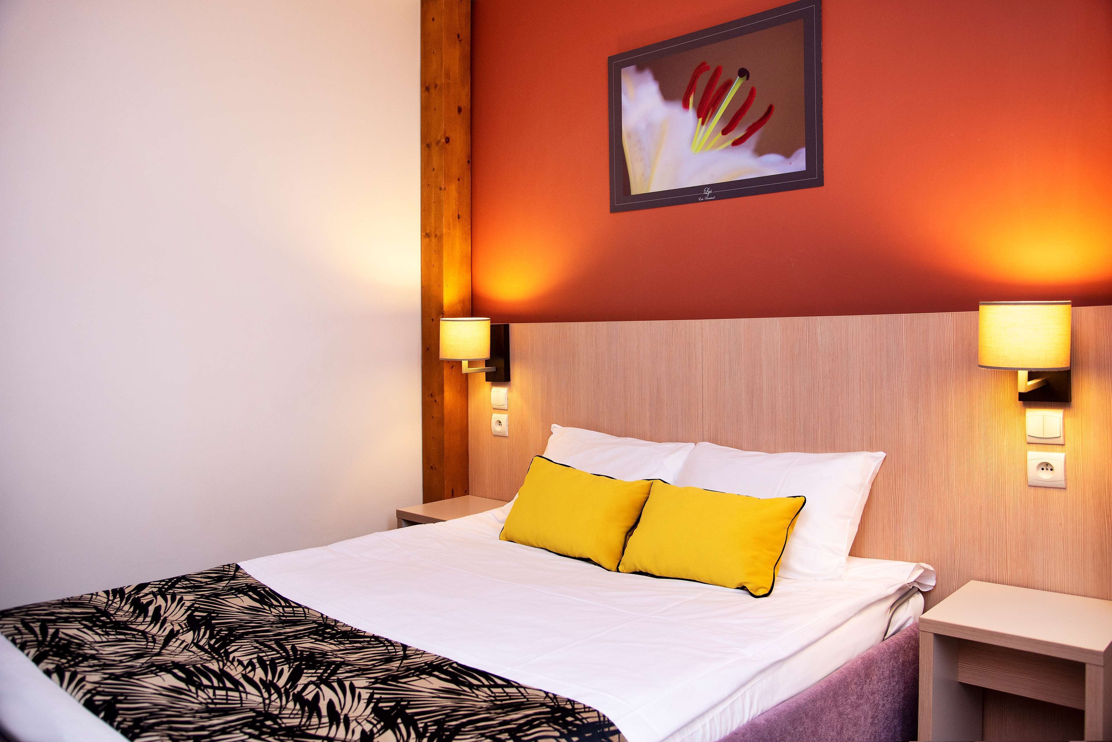 A neatly made bed with white and yellow pillows, a patterned throw, and a wooden headboard. There are two bedside lamps and a framed picture on the wall.