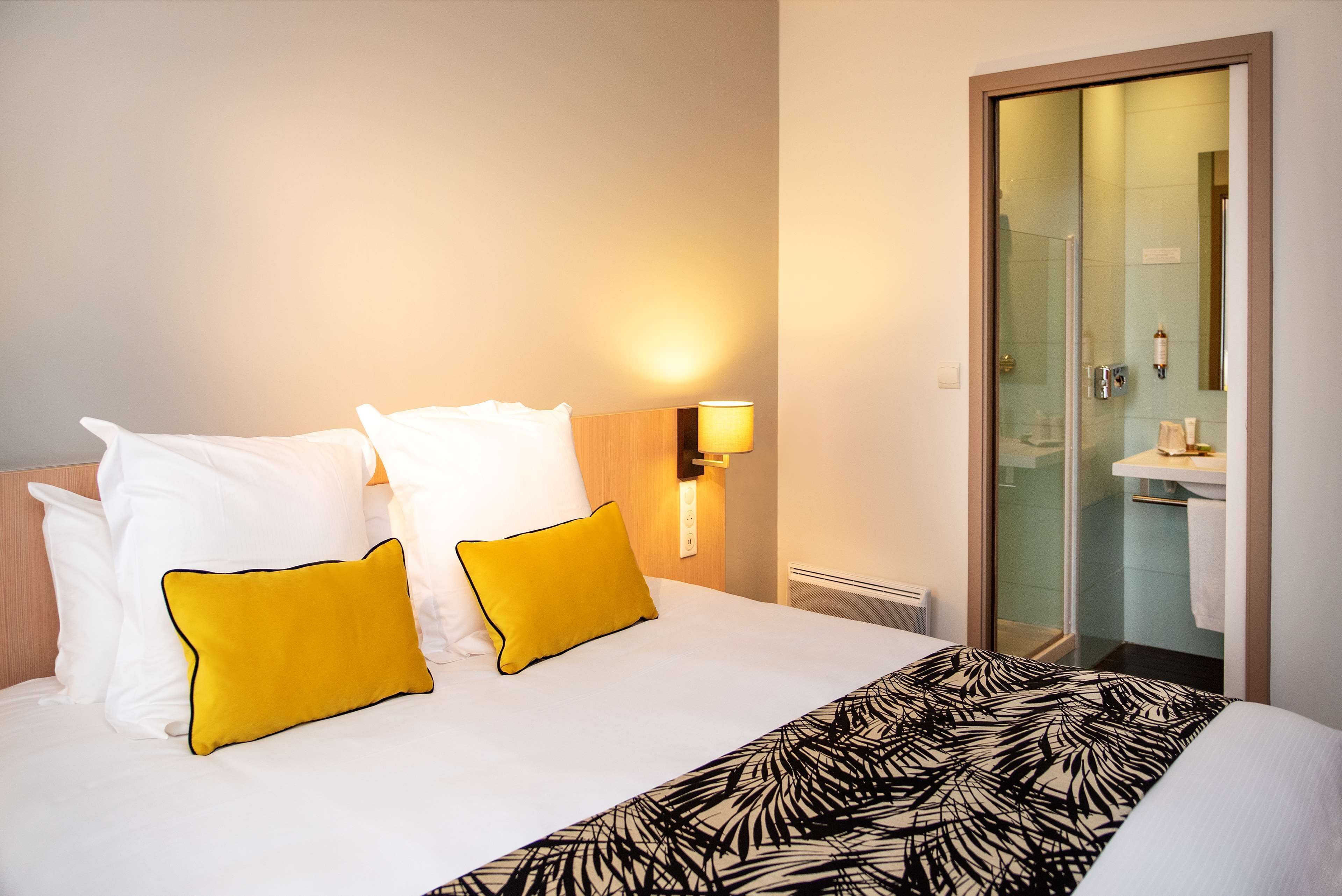 Een hotelkamer met een bed, twee kussens, een nachtkastje met een lamp, en een badkamer zichtbaar door een glazen deur.