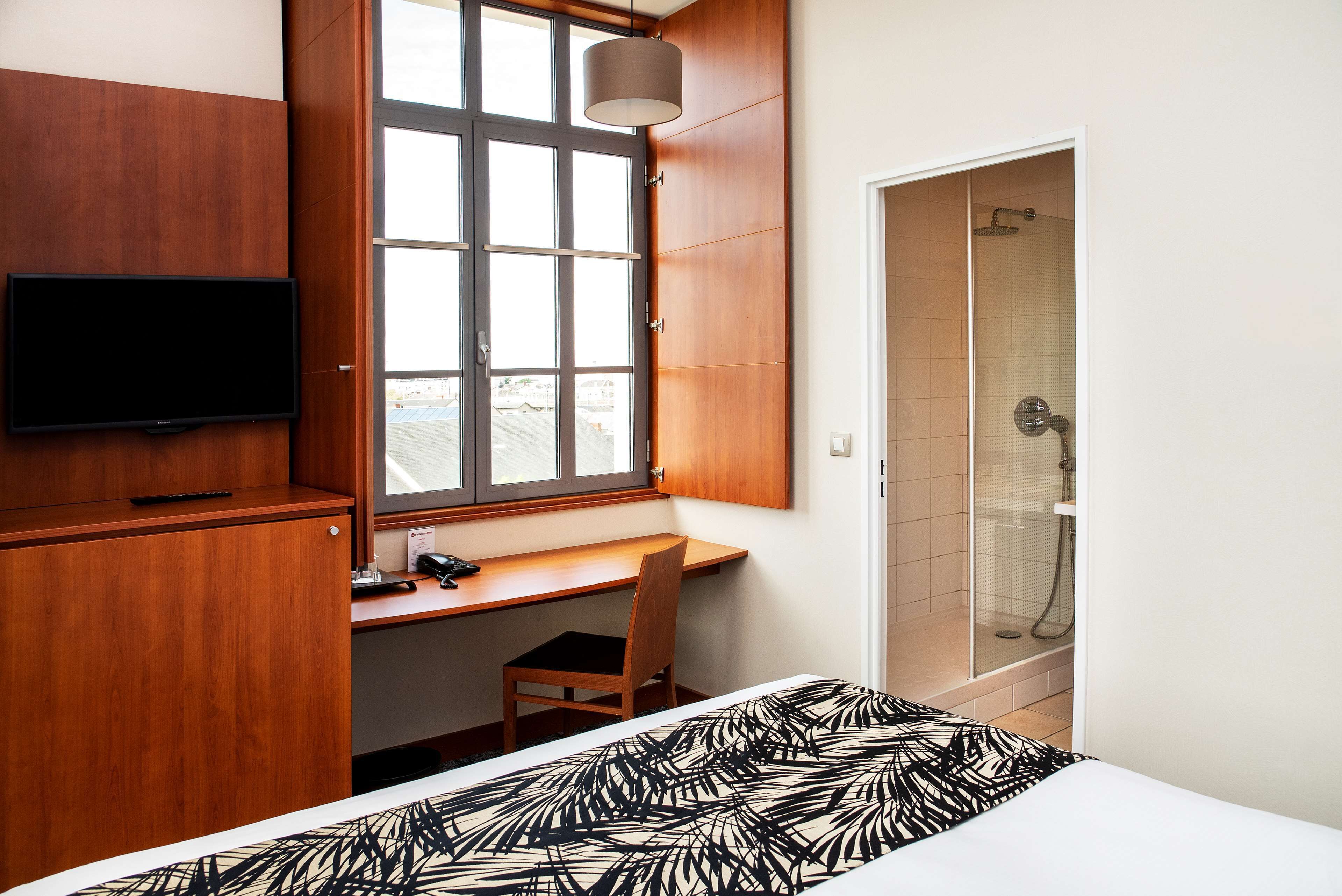 A hotel room with a wooden desk, a television, a bed with a patterned bedspread, a window with a view, and a shower area.