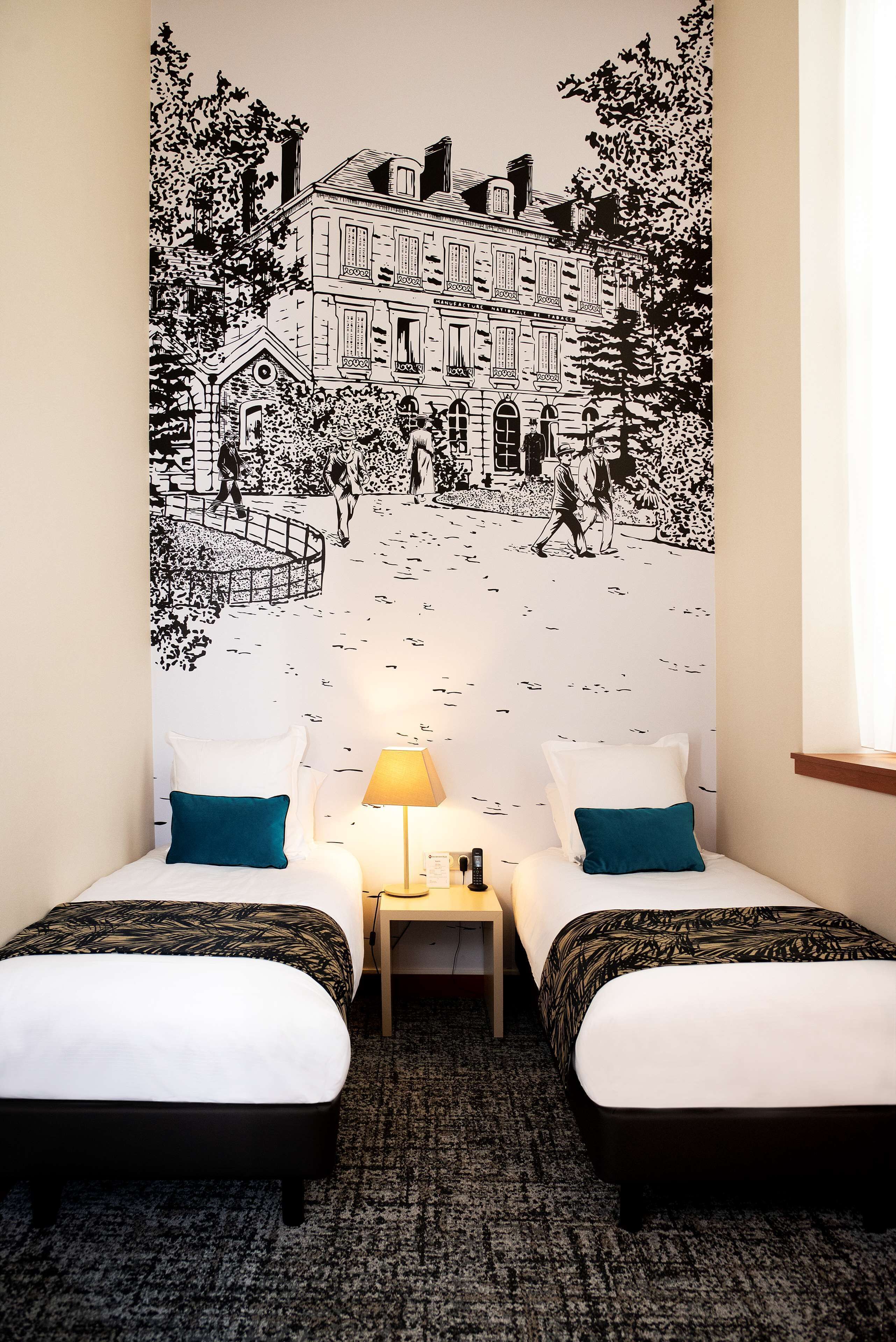 Une chambre d’hôtel avec deux lits, une table de chevet, une lampe et une grande fresque murale en noir et blanc.