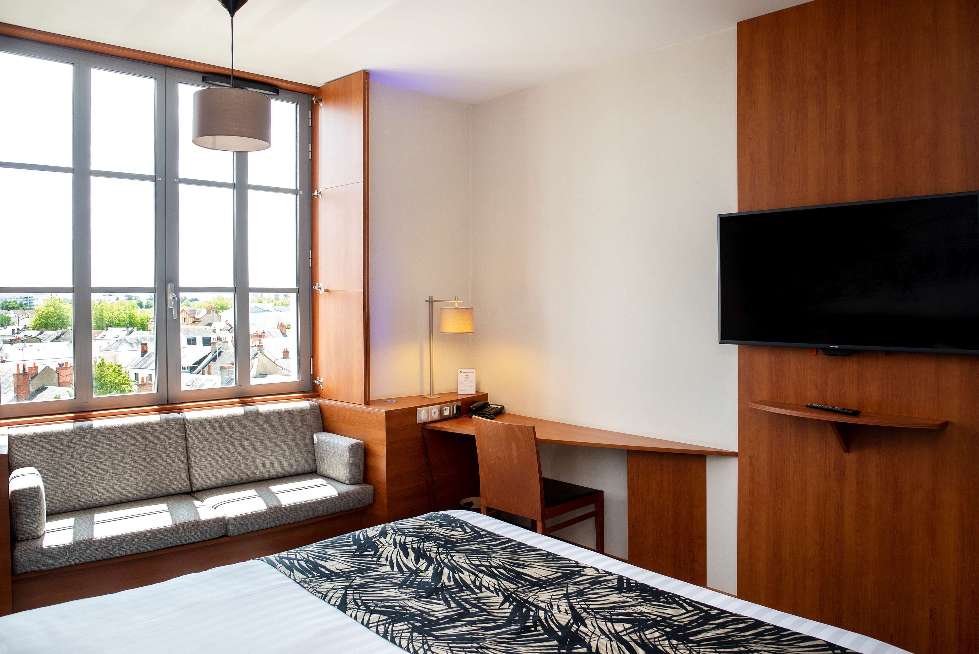 Habitación de hotel con un ventanal grande, una cama con cubrecama estampado, un escritorio con silla, una lámpara y un televisor de pantalla plana montado en la pared.