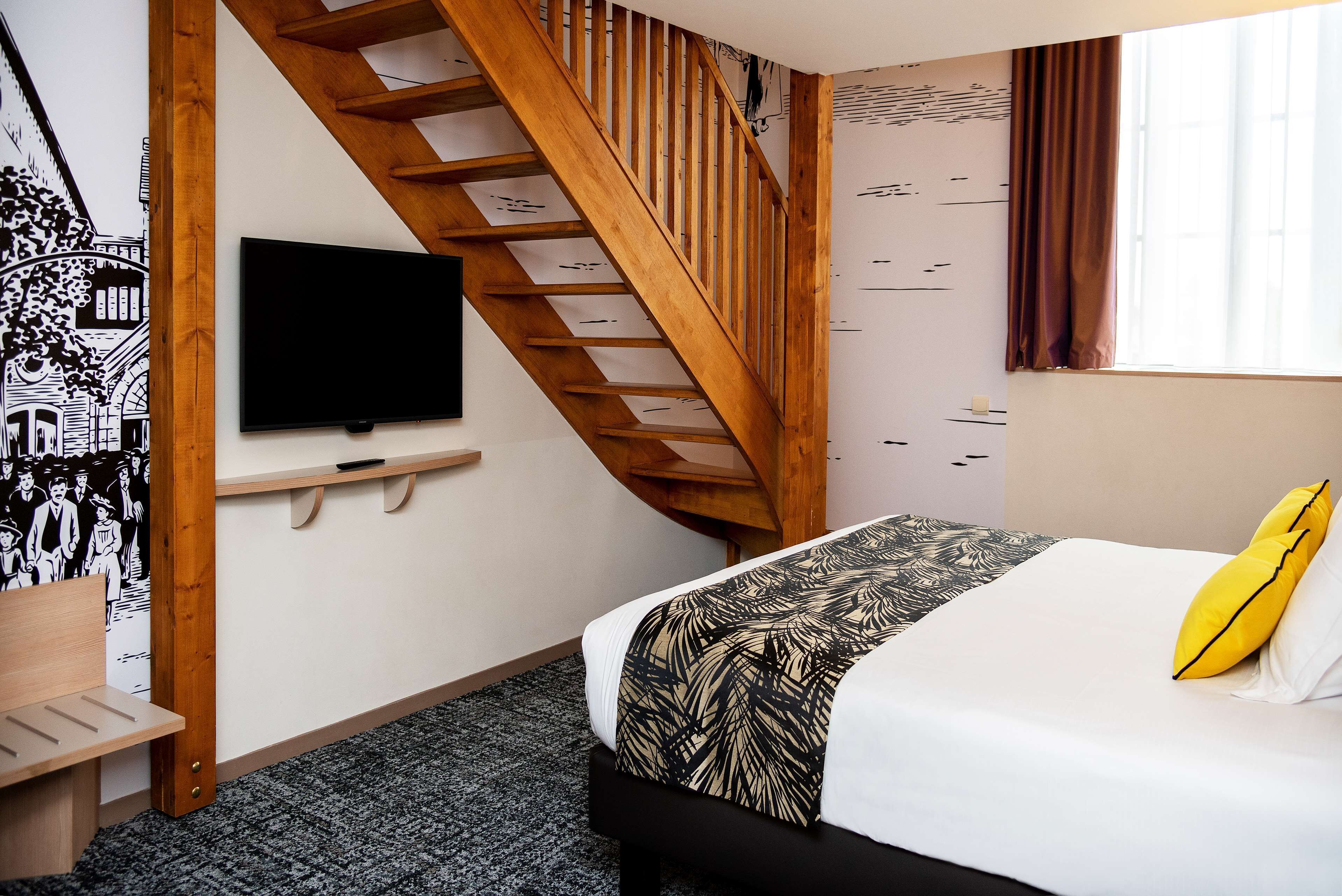 Une chambre d’hôtel avec un lit, un téléviseur fixé au mur, un escalier en bois et une fenêtre avec des rideaux.