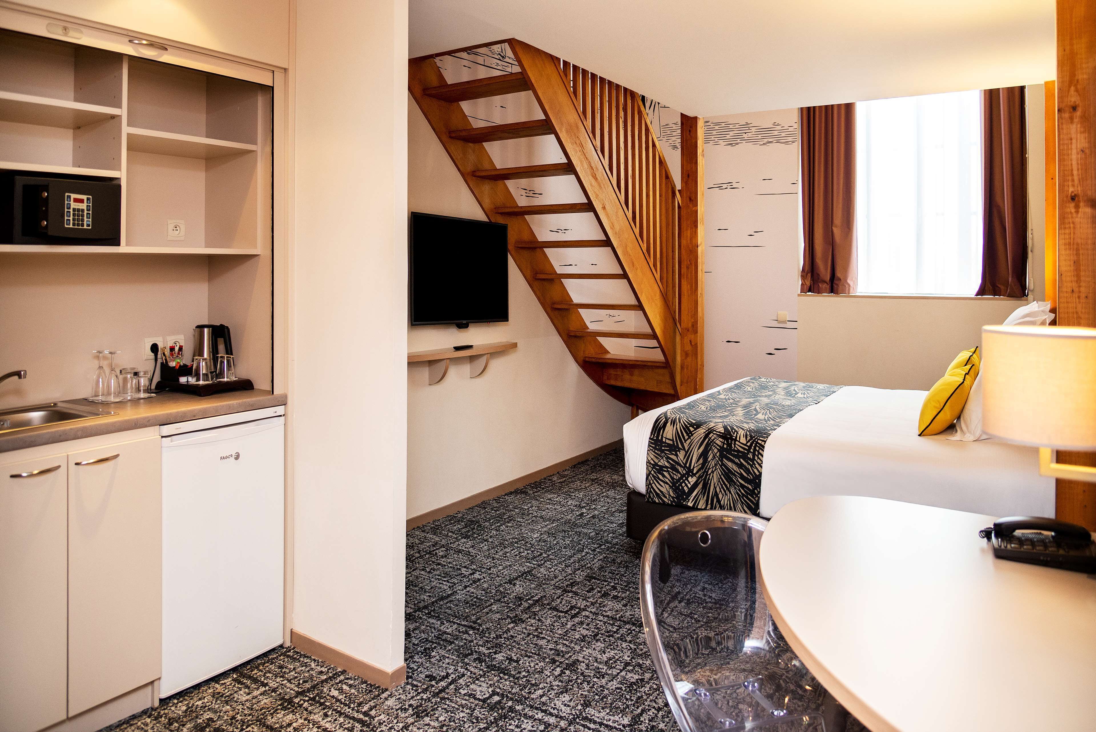 A compact hotel room with a kitchenette, a bed with a patterned bedspread, a desk with a lamp, and a staircase.