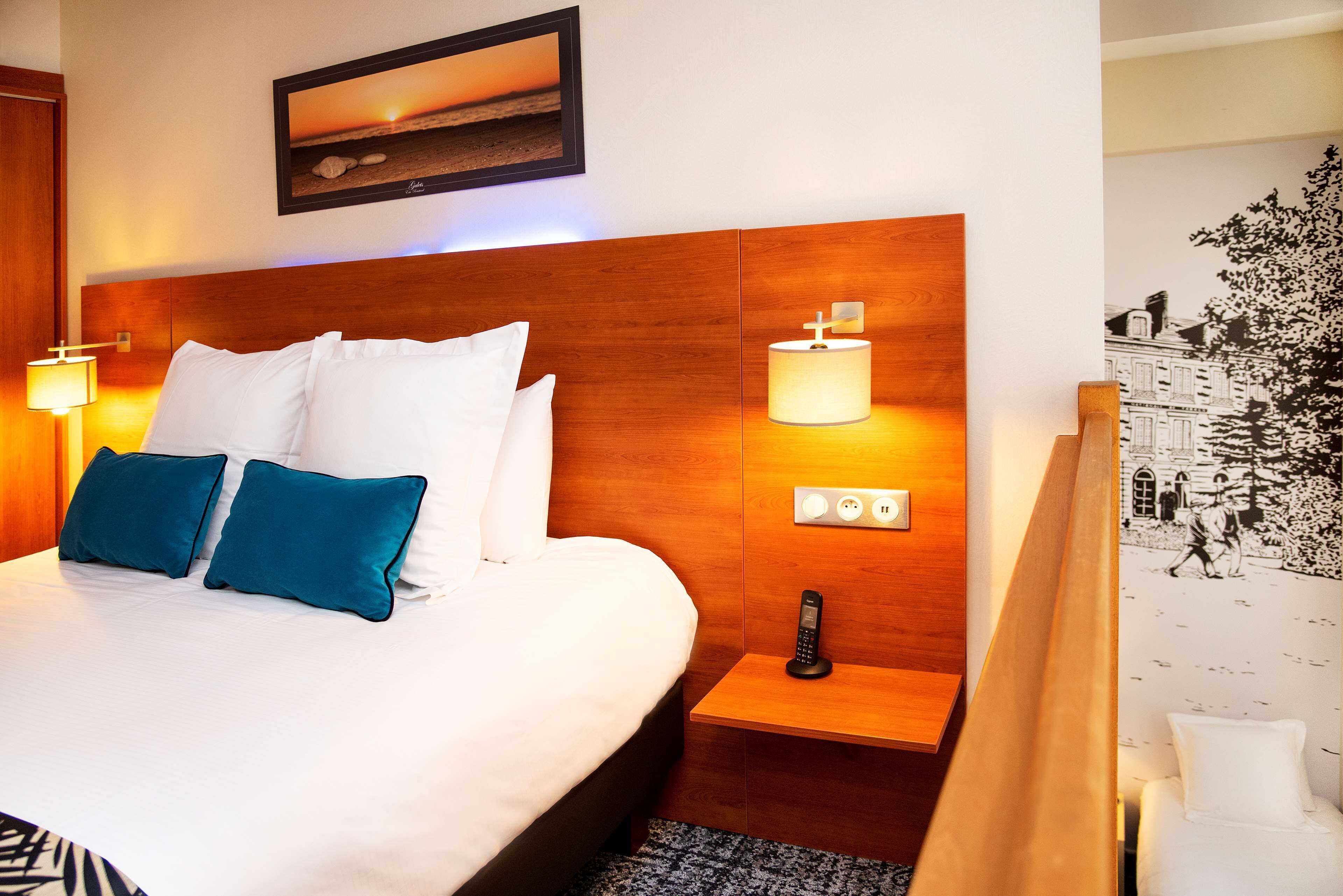 A hotel room with a large bed, two blue pillows, a bedside table with a lamp, a phone, and a wall-mounted light.