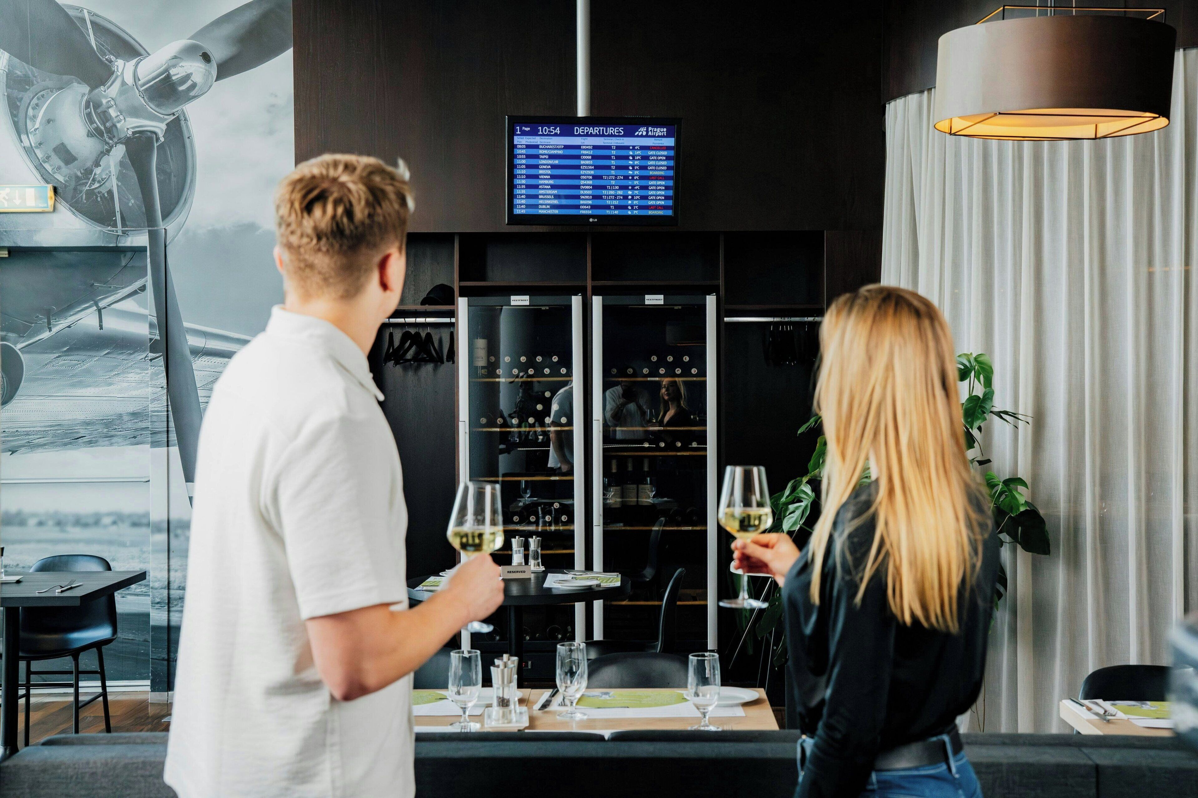 A couple toasting with wine glasses in a modern bar area with a departure board above.