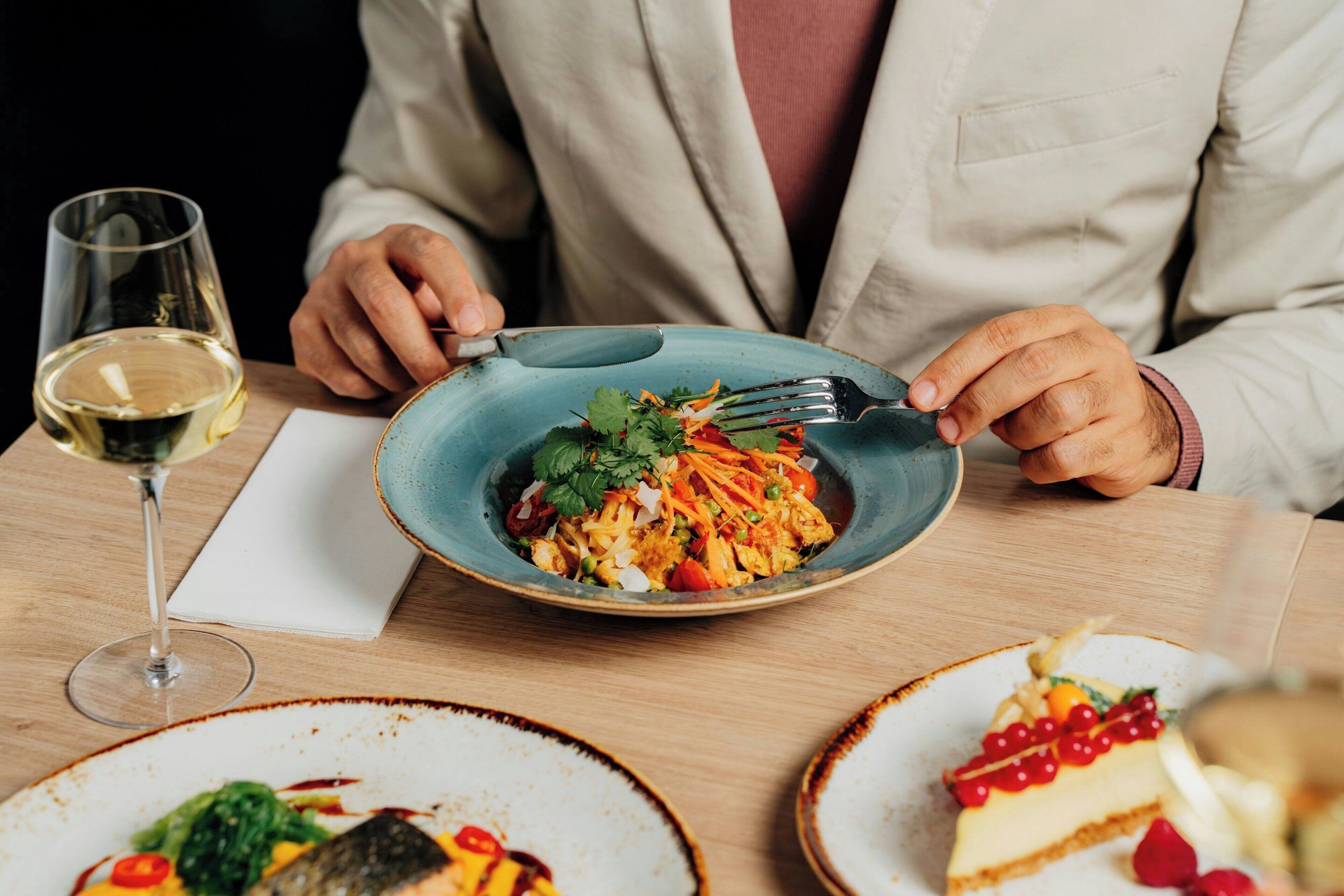 A dining table with a plate of food, a glass of wine, and a person wearing a light-colored shirt.