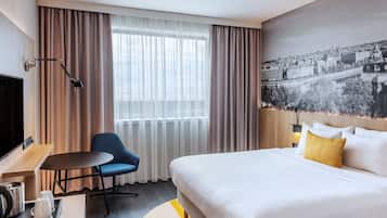 A hotel room with a bed, a desk with a kettle, a chair, a TV, and a large window with curtains.