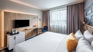 Et moderne hotelværelse med en stor seng, et skrivebord med et fladskærms-tv, en stol og et vindue med gardiner.