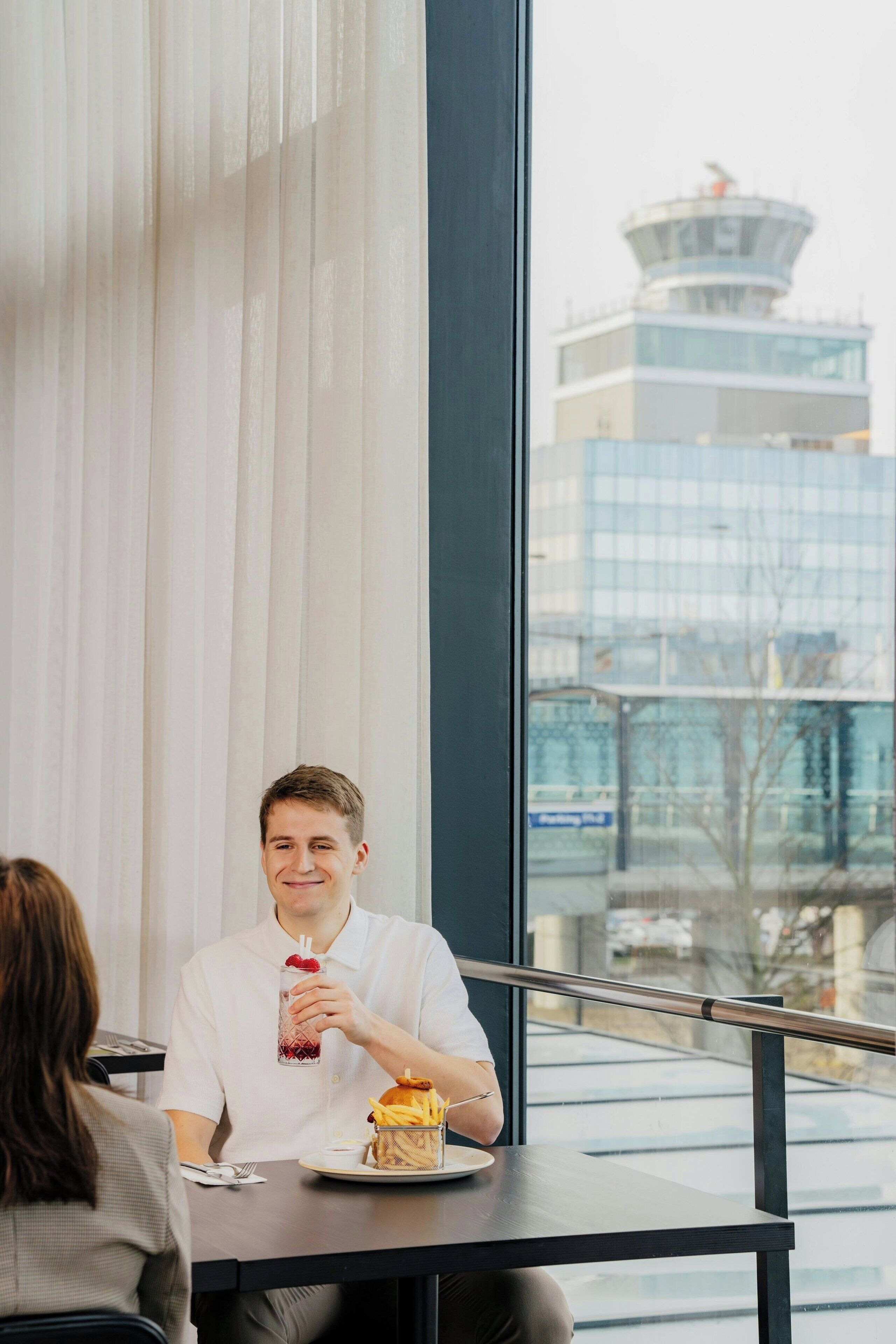 A person sitting at a table with a meal, a window with a view of a building, and a railing.