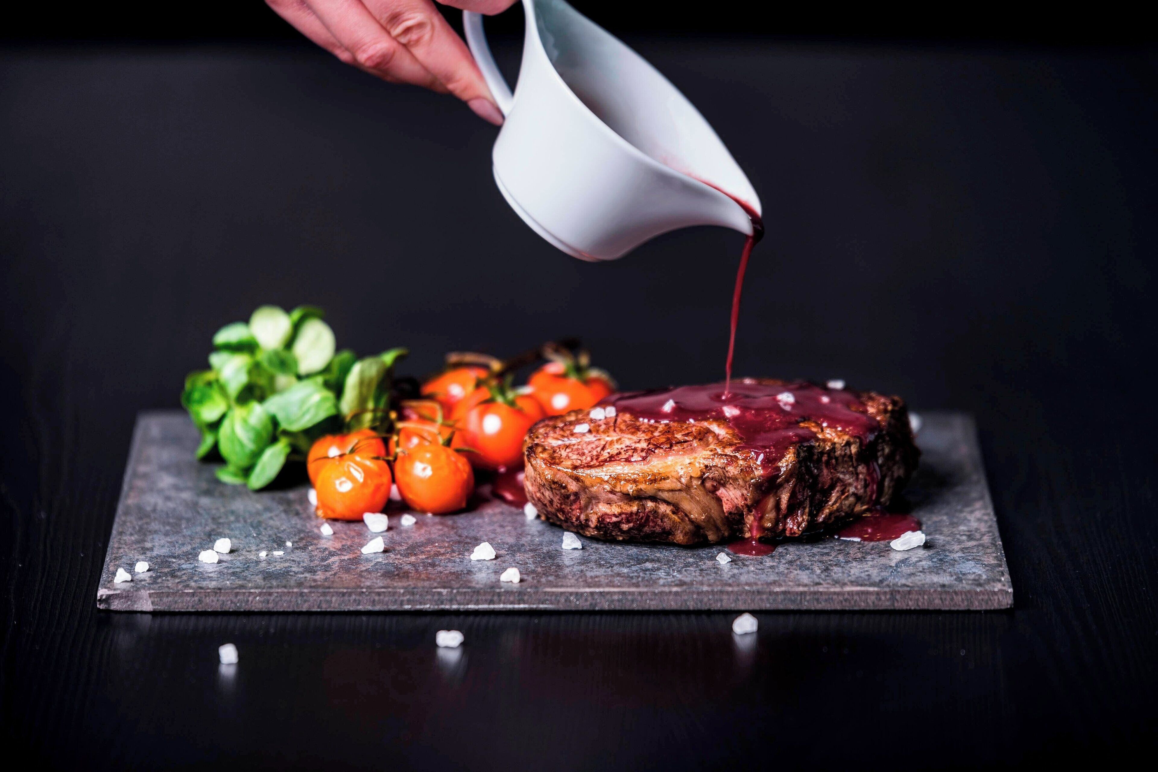 A steak with a red sauce being poured on it, surrounded by tomatoes and basil leaves on a dark slate board.