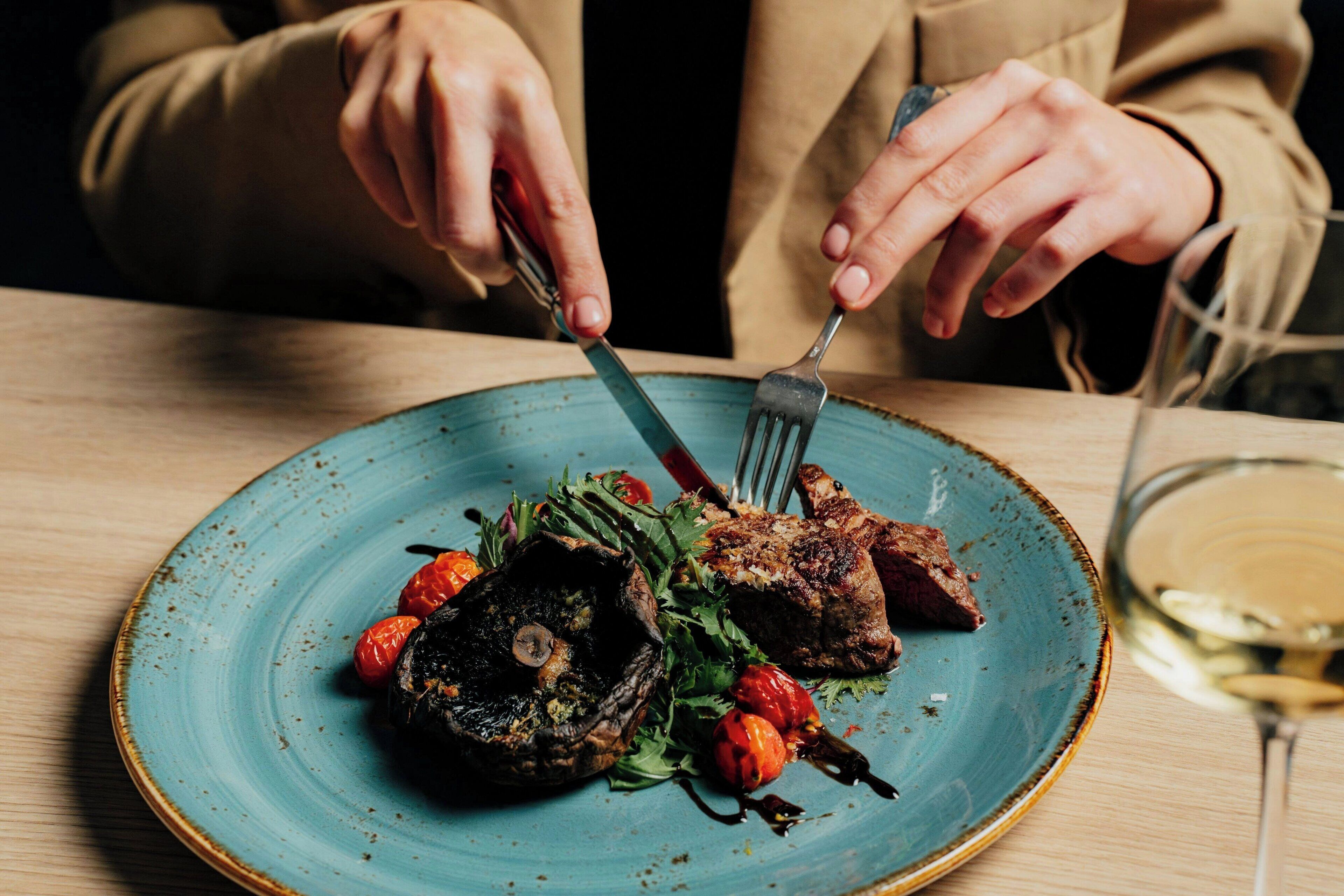 A person holding a fork and knife over a plate of food with a glass of white wine.