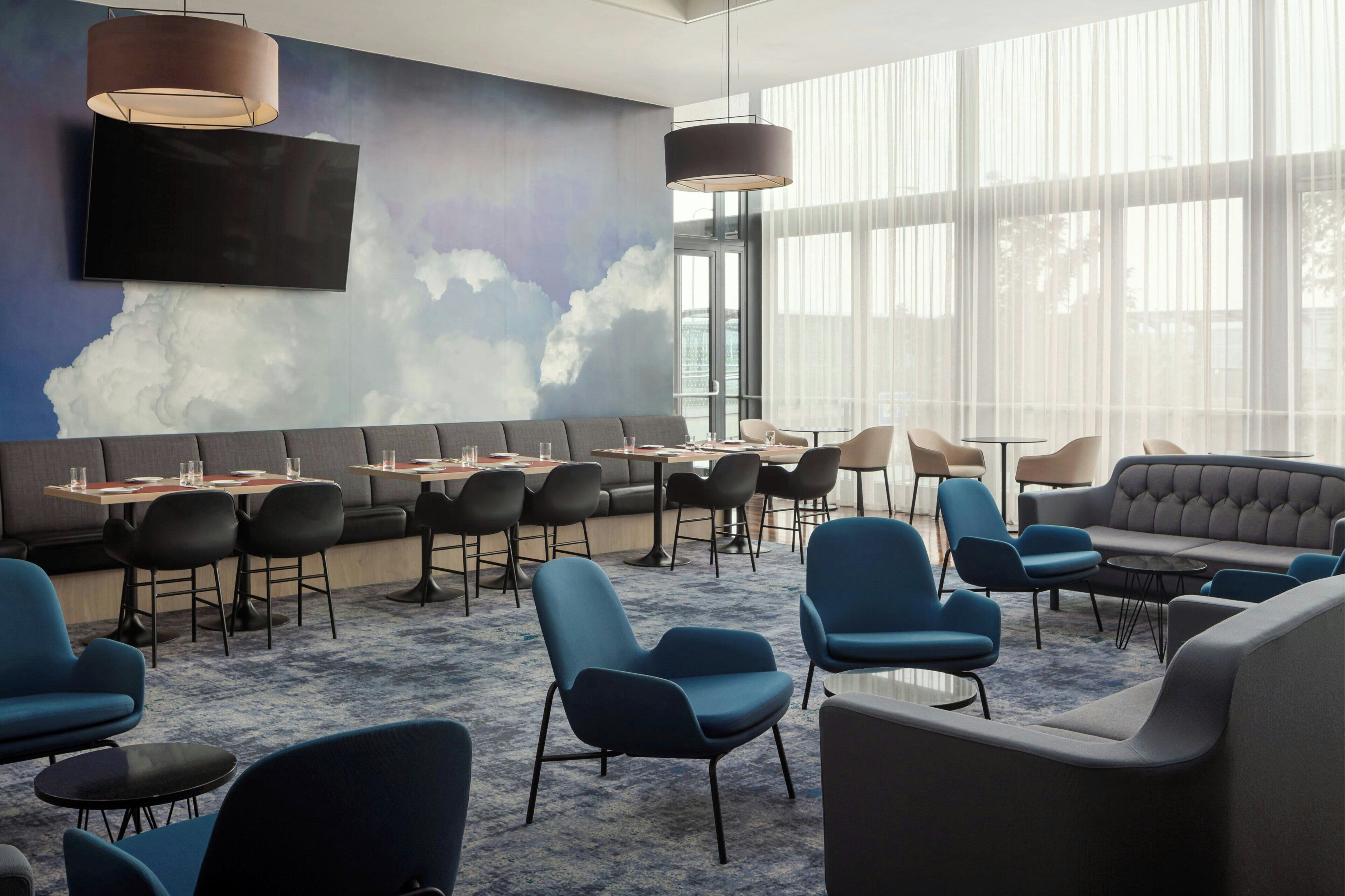 Un área de lounge moderna con una pared con motivos de nubes, diversas opciones de asientos que incluyen sofás y sillones, y grandes ventanales que ofrecen luz natural.