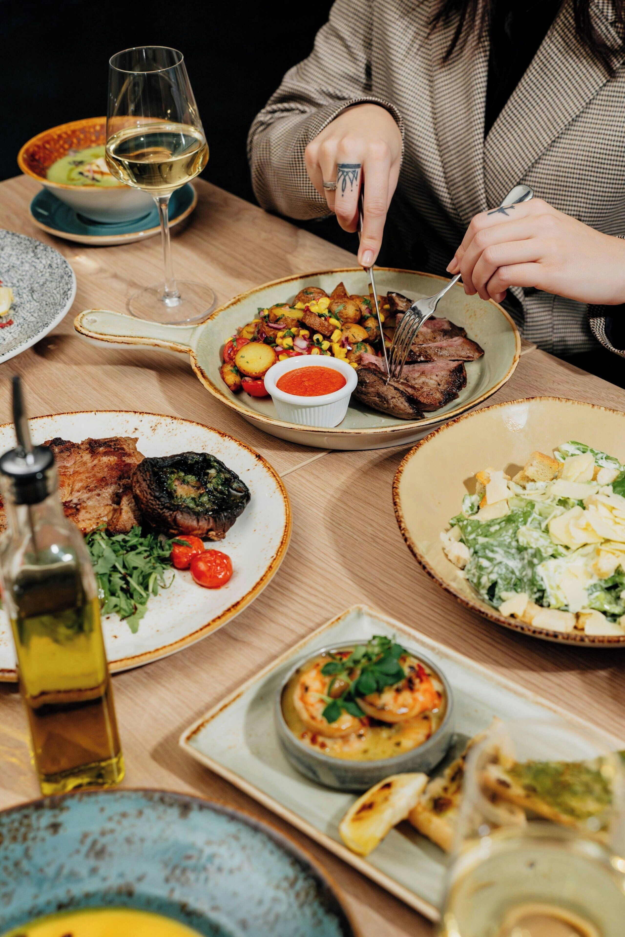 A table with various dishes including steak, salad, and shrimp, along with a glass of white wine.