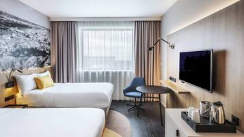 A hotel room with two beds, a TV, a desk with coffee-making equipment, and a large window with curtains.