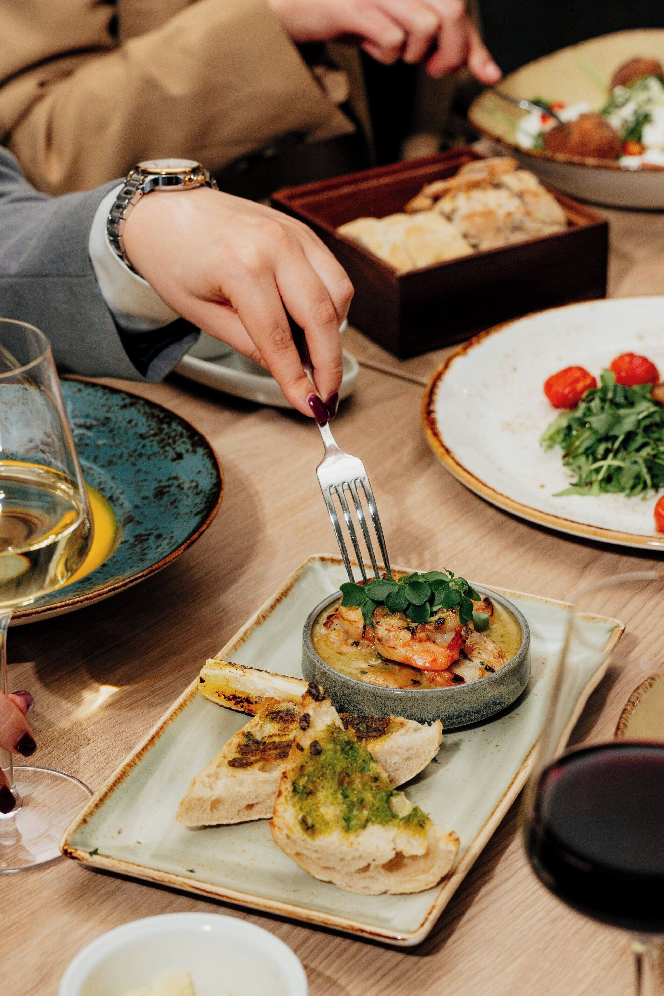 Une table à manger avec une assiette de nourriture, un verre de vin blanc et une personne qui utilise une fourchette.