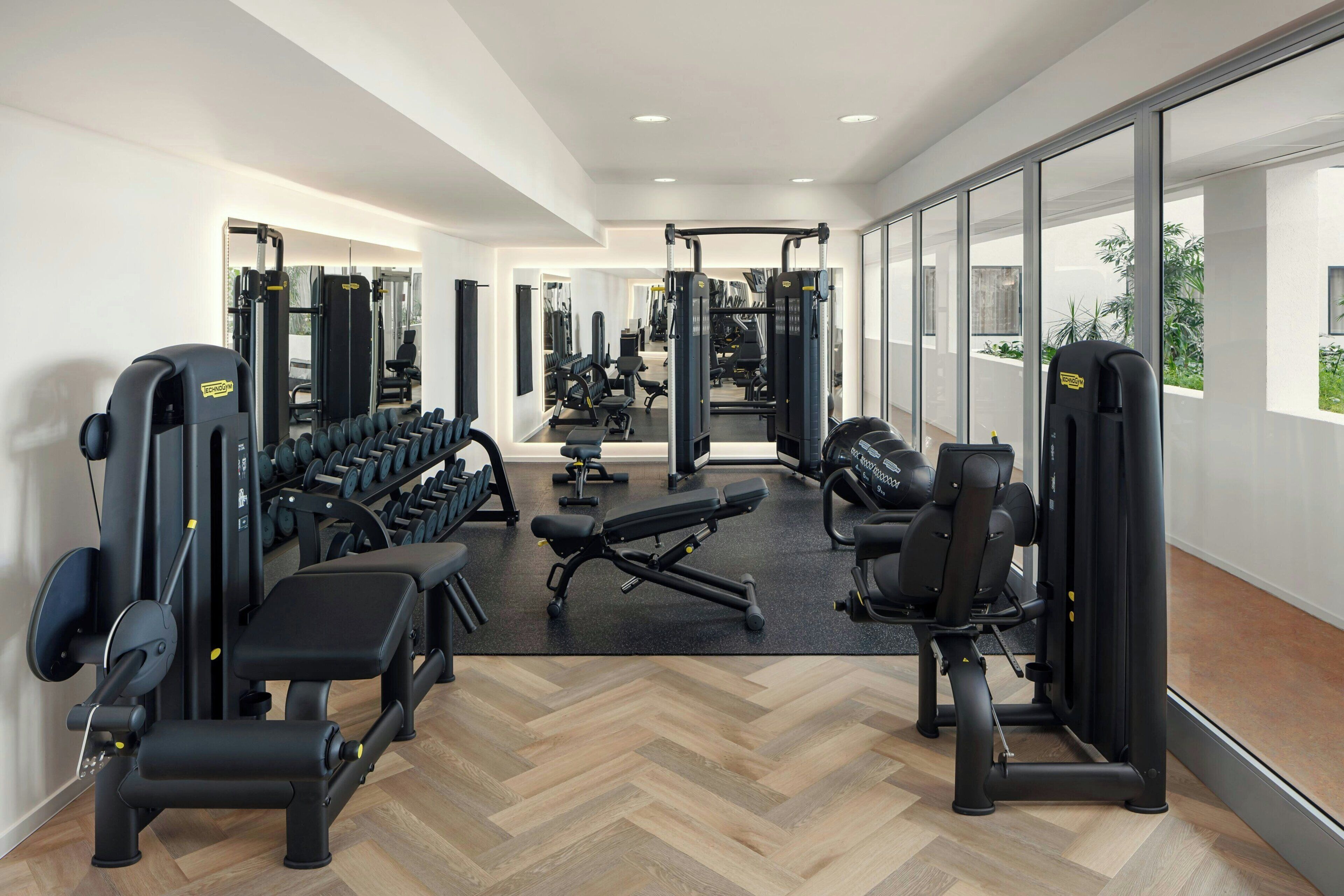 A modern gym with various exercise equipment, mirrored walls, and large windows.