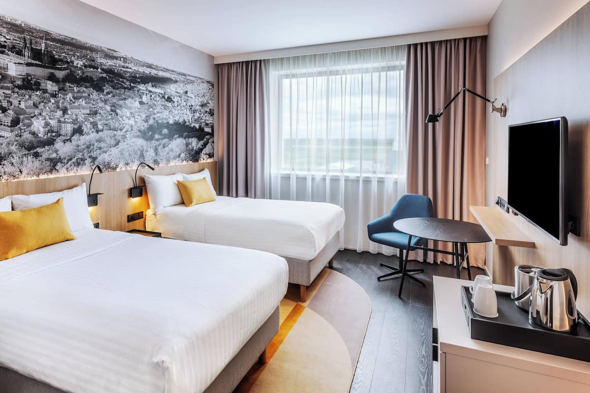 Een hotelkamer met twee bedden, een bureau met waterkoker, een stoel, een televisie en een groot raam met gordijnen.