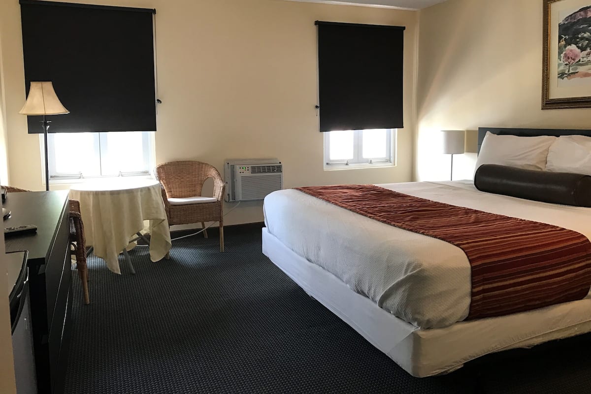 A hotel room with a bed, a chair, a small table, and a window with blinds.