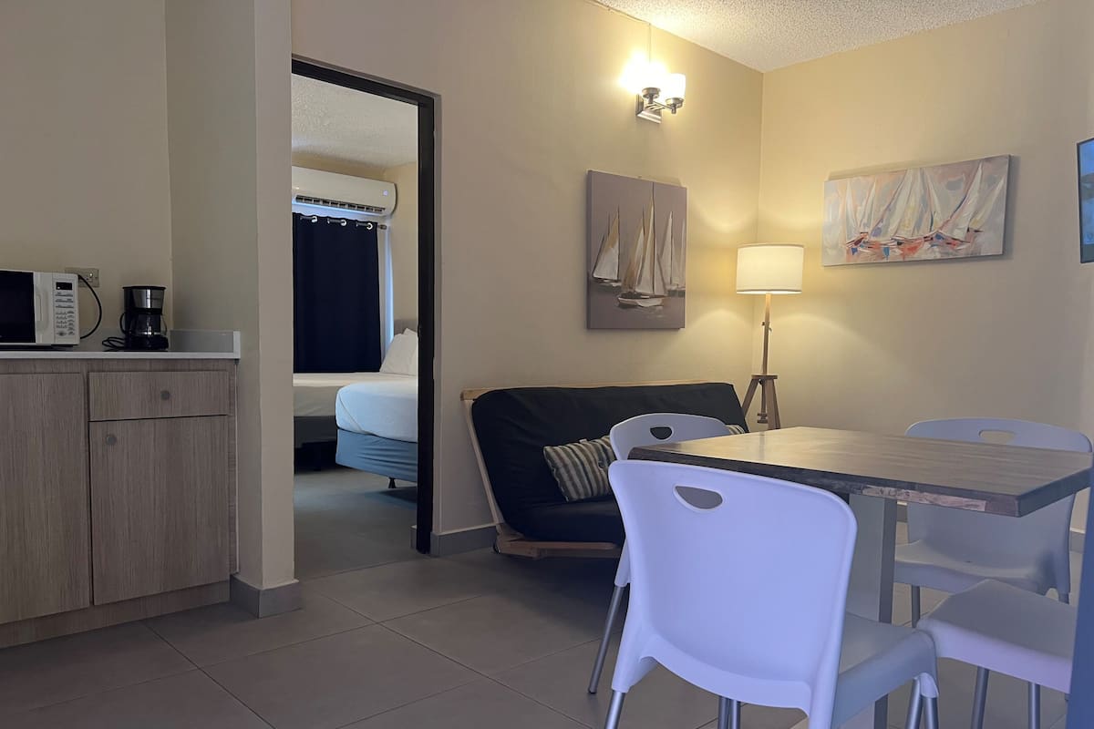 A compact hotel room with a kitchenette, a bed, a dining area, and a small living space.