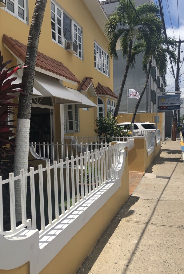 A yellow building with a white fence, palm trees, and a parked car.