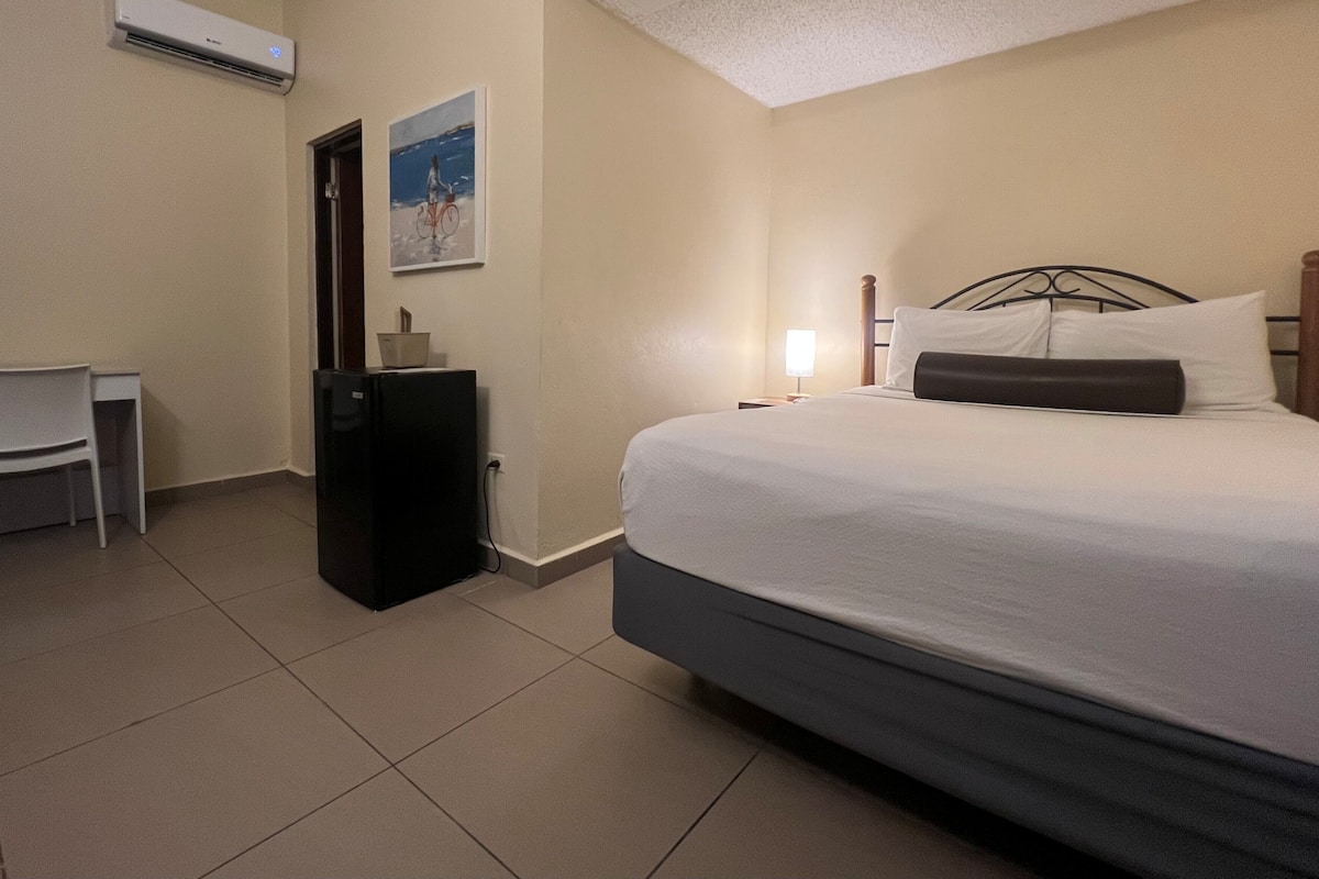 A hotel room with a bed, a desk, a chair, a refrigerator, and a wall-mounted air conditioner.
