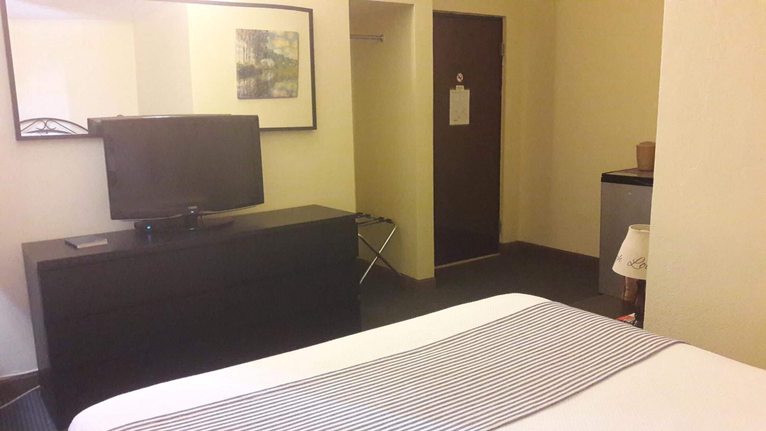 A hotel room with a bed, a television on a dresser, a door, and a small table with a lamp.