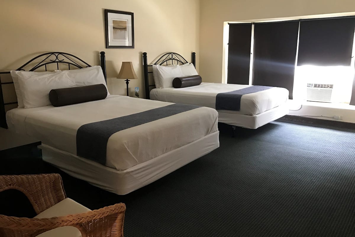 A hotel room with two beds, a large window with blinds, a framed picture on the wall, and a wicker chair.