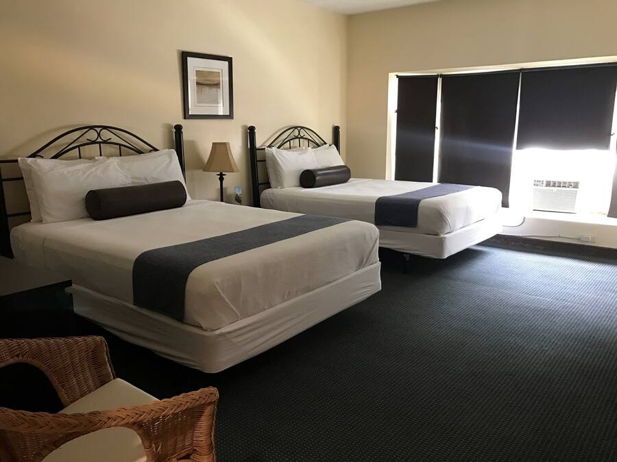 A hotel room with two beds, a large window with blinds, a framed picture on the wall, and a wicker chair.