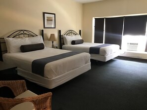 A hotel room with two beds, a large window with blinds, a framed picture on the wall, and a wicker chair.